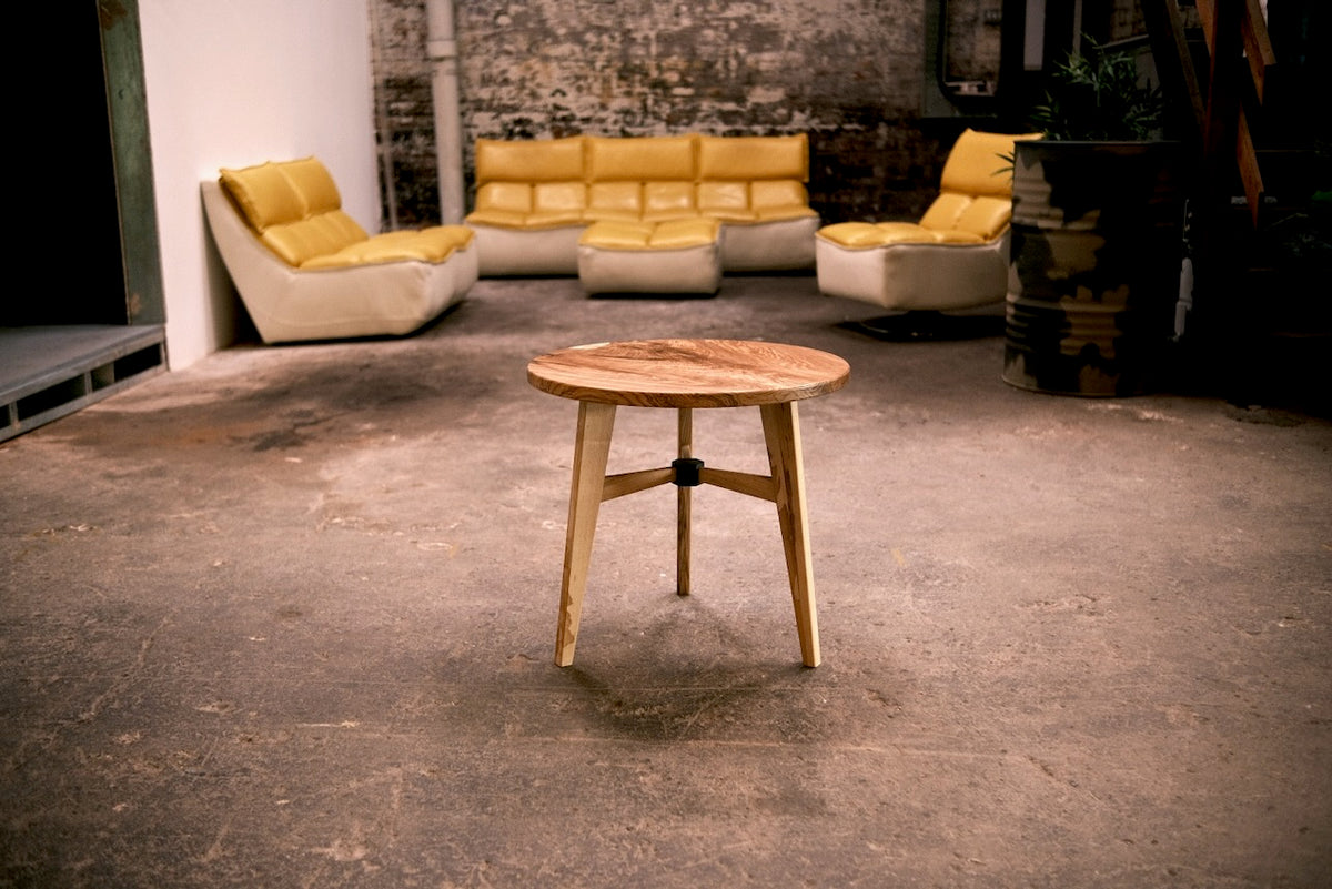 A round wooden side table with three legs stands in the foreground of a loft-style room with concrete floors, a brick wall, and modern yellow and cream seating in the background.