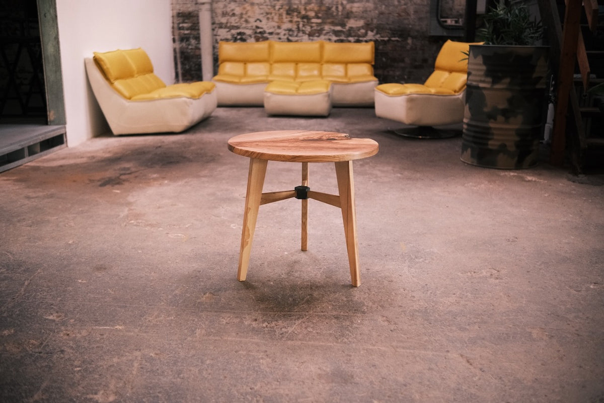 A round wooden side table with three legs and a natural wood top, set on a concrete floor in a loft-style room with yellow sofas in the background.