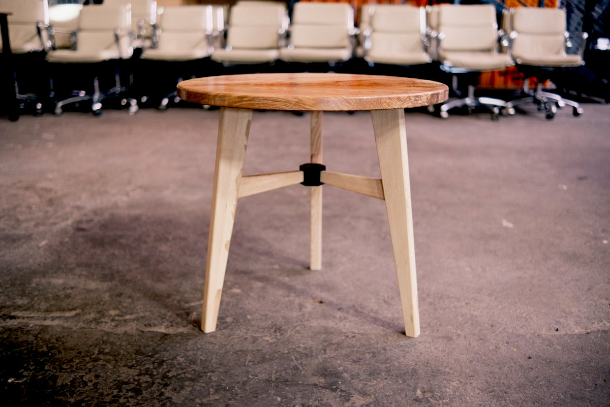 A round wooden table with a light natural ash wood top and three angled legs, featuring a central black support, set in an indoor space with a concrete floor and office chairs in the background.