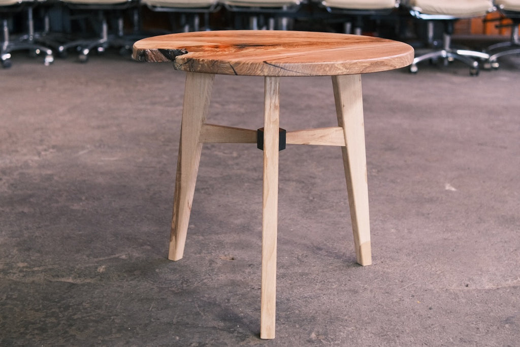 A stylish wooden side table with a live-edge round top and three tapered legs, shown on a concrete floor in an indoor setting with office chairs blurred in the background.