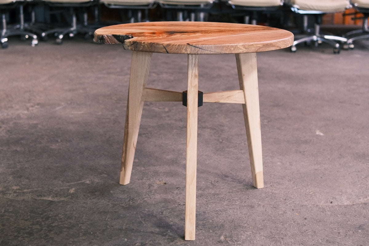 A stylish wooden side table with a live-edge round top and three tapered legs, shown on a concrete floor in an indoor setting with office chairs blurred in the background.