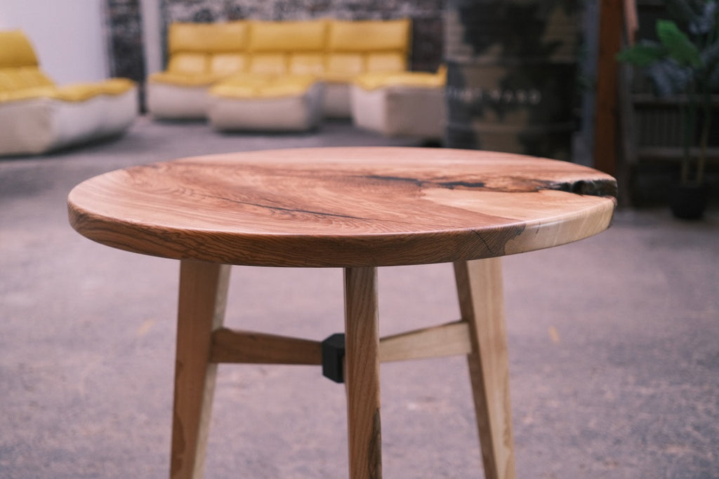A round wooden side table with a live-edge top and three angled legs, shown in a blurred indoor setting with yellow seating in the background.