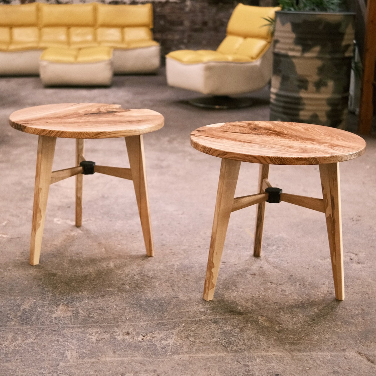 Two round wooden side tables with natural wood tops and angled legs, shown in an industrial setting with a concrete floor and retro lounge furniture in the background.