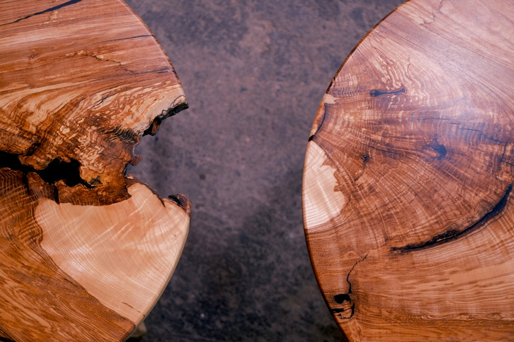 Close-up of two round ash table tops with natural live edges and rich wood grain, set against a dark textured surface.