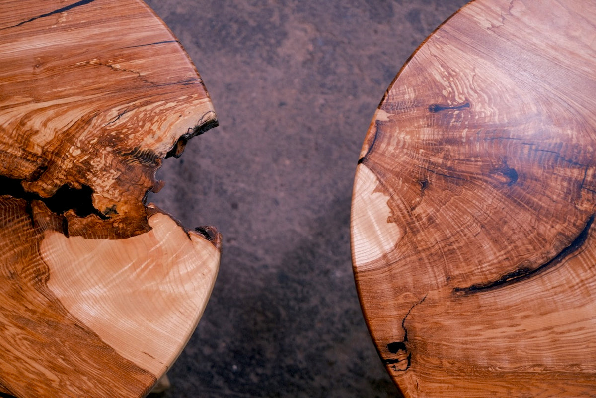 Close-up of two round ash table tops with natural live edges and rich wood grain, set against a dark textured surface.