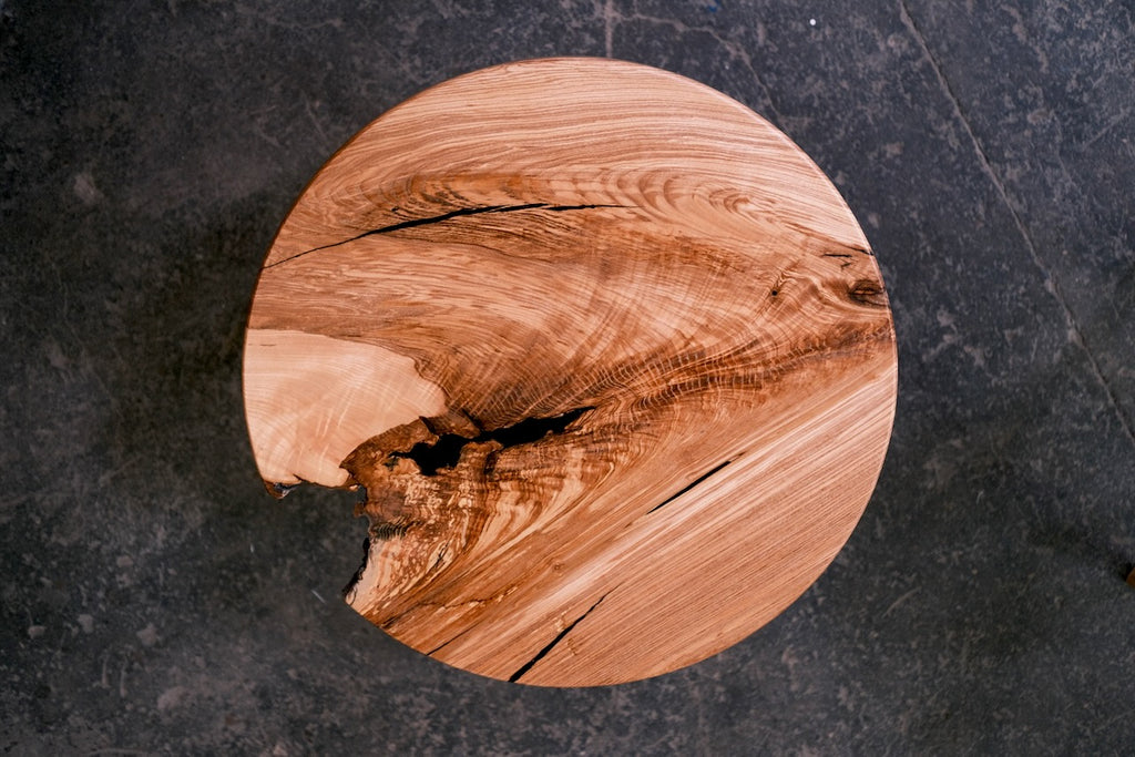 A round ash table with a natural live edge and rich wood grain, displayed on a dark textured slate surface.
