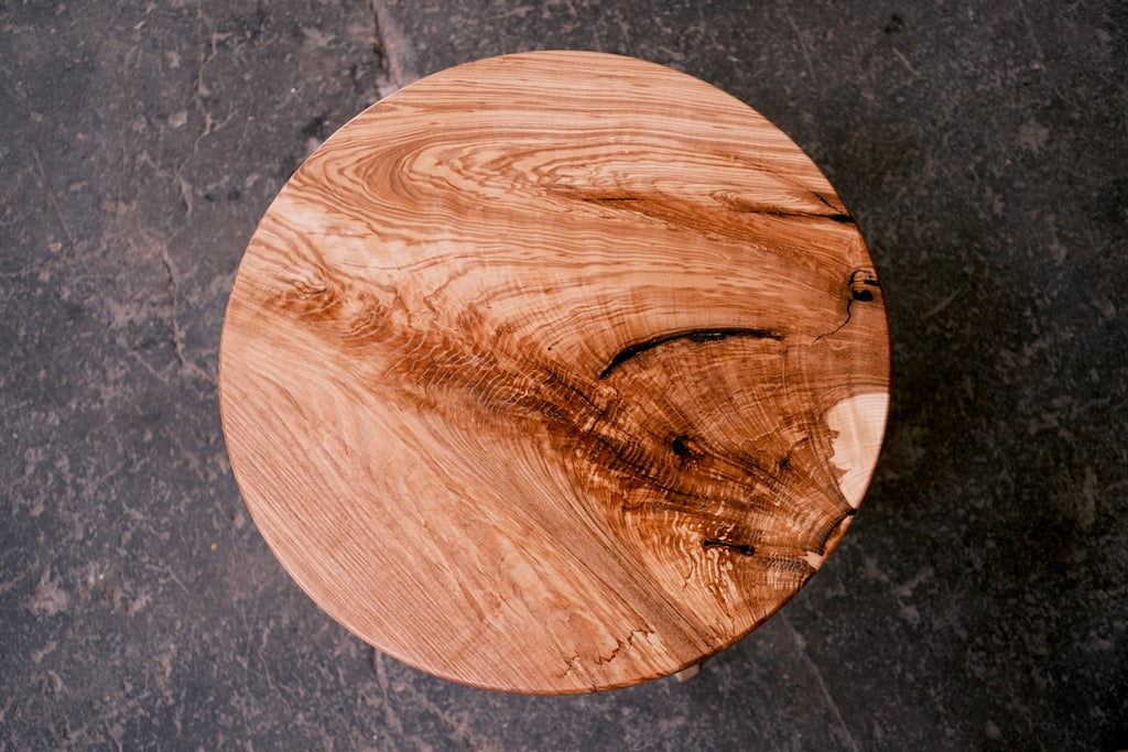 A round ash table with a natural live edge and rich wood grain, displayed on a dark textured slate surface.