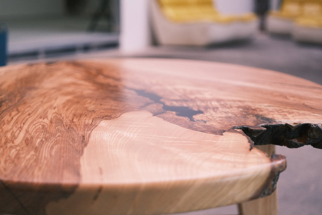 A close-up of a round live-edge wooden table with a polished finish, showcasing natural wood grain patterns and a dark resin inlay, set against a blurred indoor background.