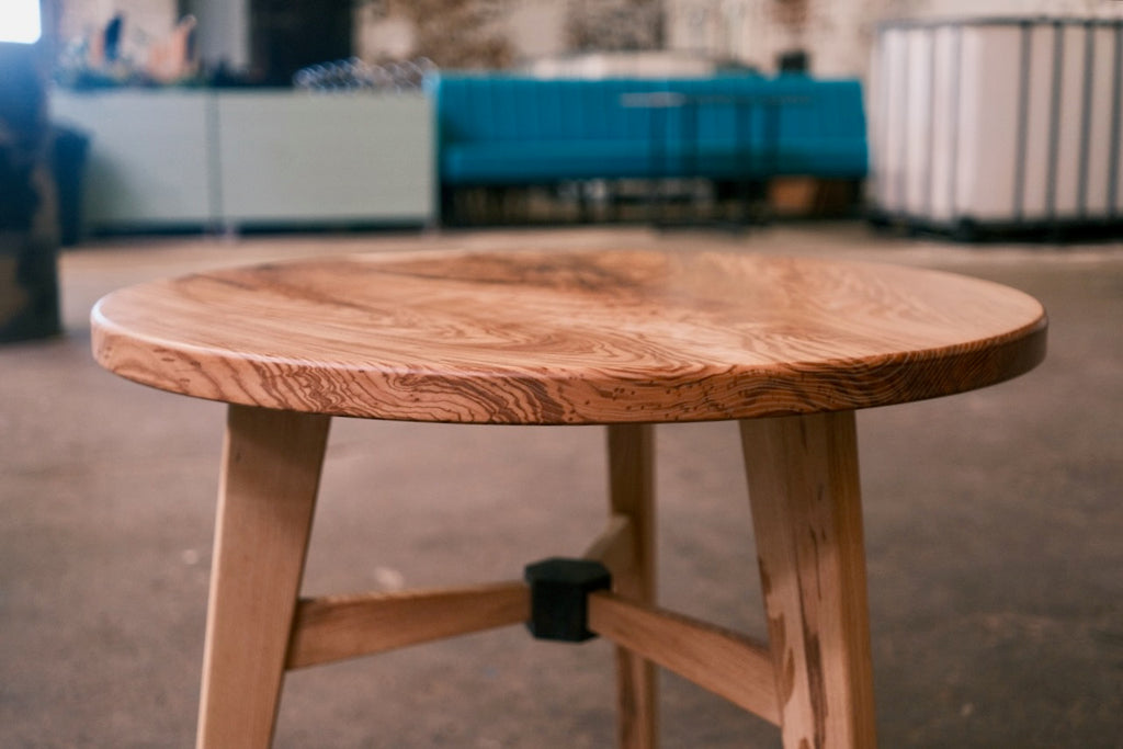A close-up of a round wooden table with a natural ash top and light-colored legs, set in a workshop or studio environment with blurred furniture in the background.