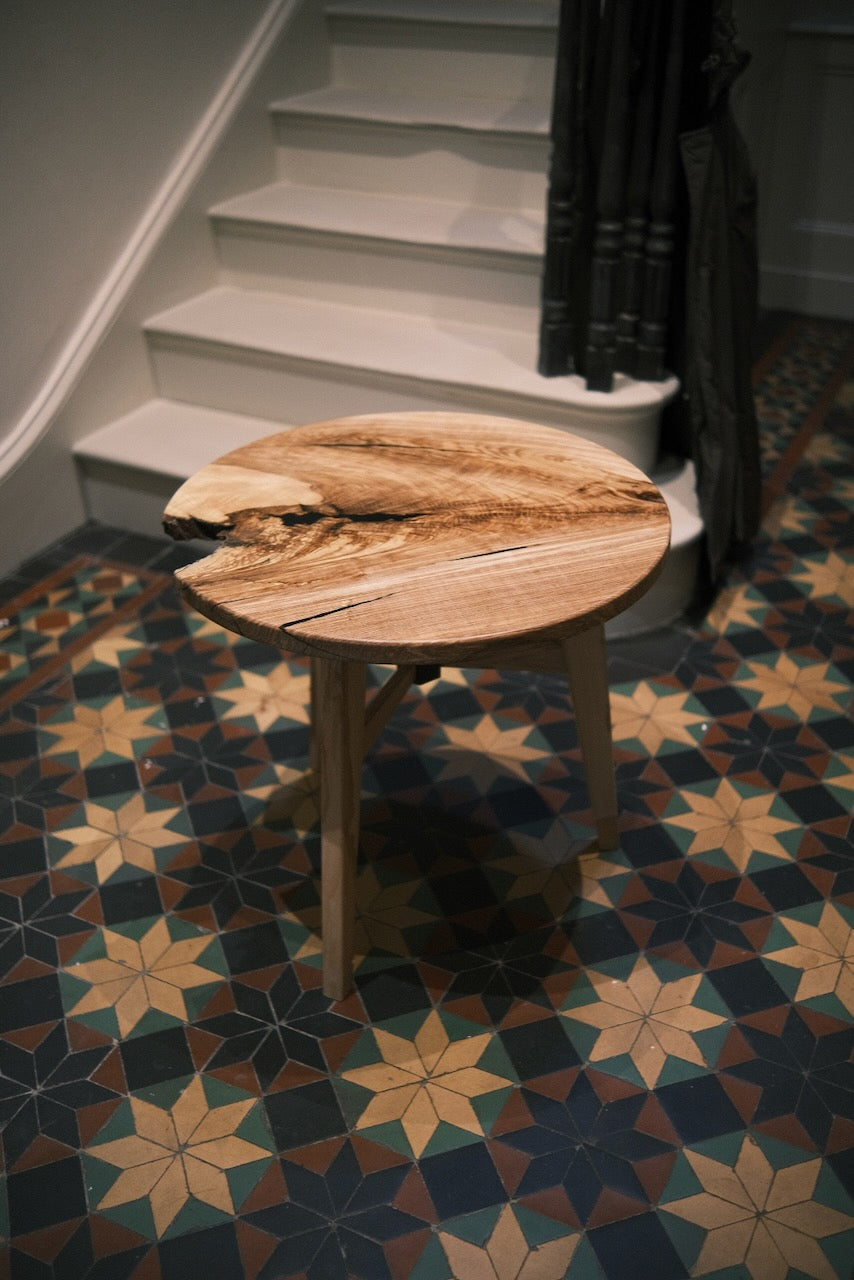 A round ash wooden table with a live-edge natural wood top and three legs, placed on a patterned geometric tile floor in front of white stairs.