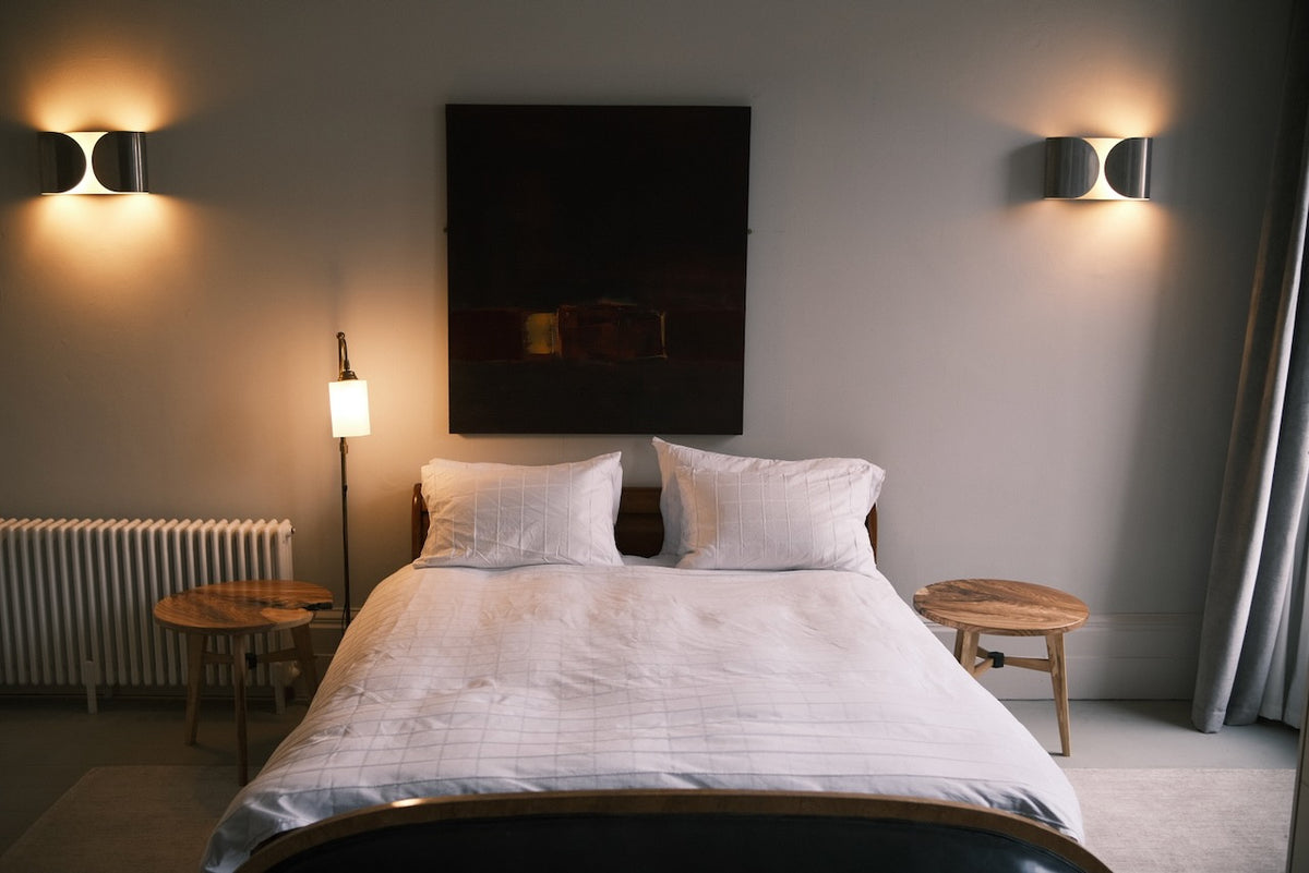A modern bedroom featuring a neatly made bed with white bedding, a dark rectangular wall art piece, two wall-mounted sconces, a floor lamp, and two small ash wooden side tables.