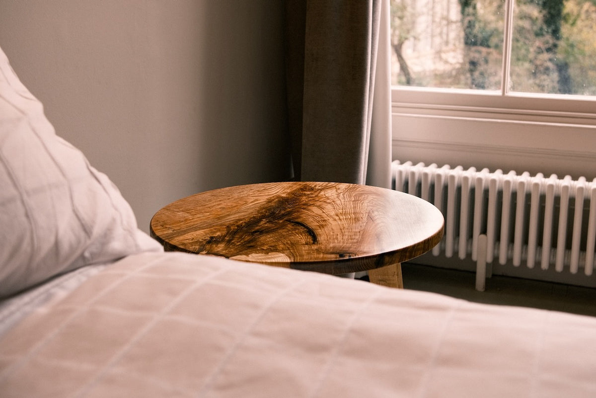 A round wooden nightstand with a live-edge natural wood top, positioned next to a bed with white bedding in a softly lit bedroom.