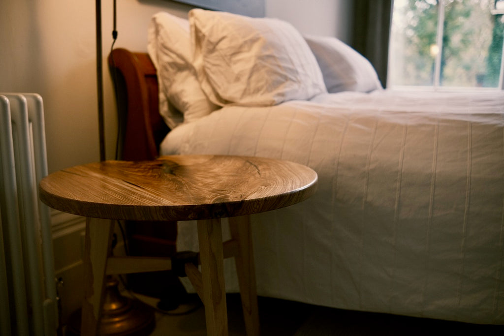 A rustic round wooden nightstand in a bedroom setting next to a bed with white bedding and pillows.
