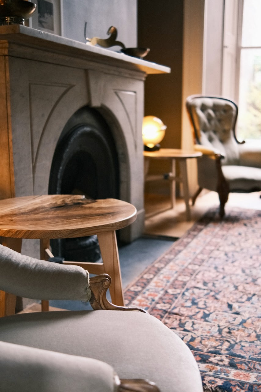 A cozy interior scene featuring a light beige upholstered armchair in the foreground, a round wooden side table, a stone fireplace, and a patterned area rug. A second tufted armchair sits near a window in the softly blurred background.