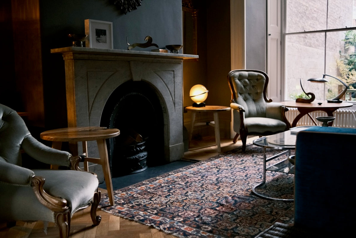 A styled interior room scene featuring multiple upholstered armchairs, a stone fireplace, a patterned area rug, side tables, and warm ambient lighting near a large window.