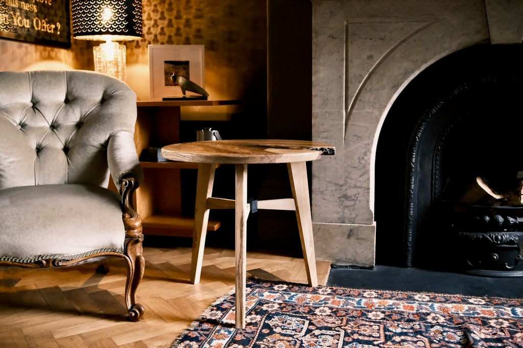 A round wooden side table with a live-edge natural top, positioned in a cozy, dimly lit living room next to a tufted armchair and in front of a stone fireplace.