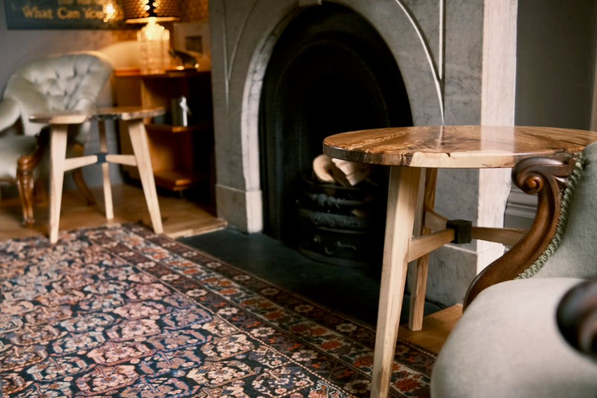 A rustic live-edge wooden side table in a cozy, dimly lit room with a stone fireplace, patterned rug, and upholstered chairs.