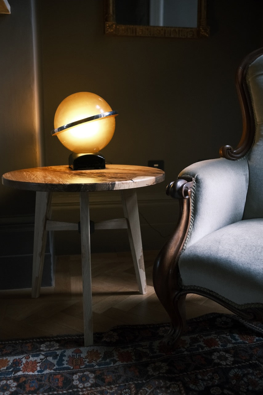 A glowing globe-style table lamp with a black base sits on a round wooden side table next to an upholstered armchair in a dimly lit room.