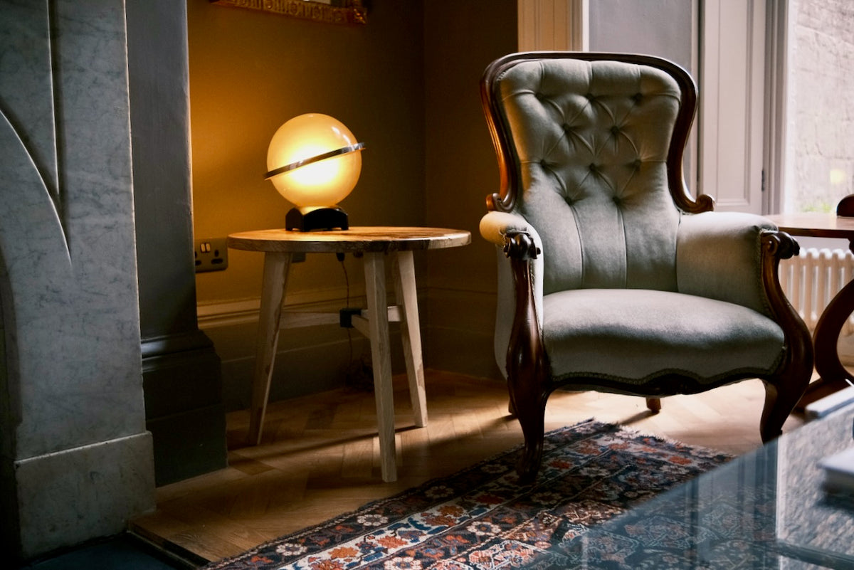 Solid ash wooden table with a glowing globe lamp next to a classic armchair with dark wood arms and legs, positioned in a warmly lit room.