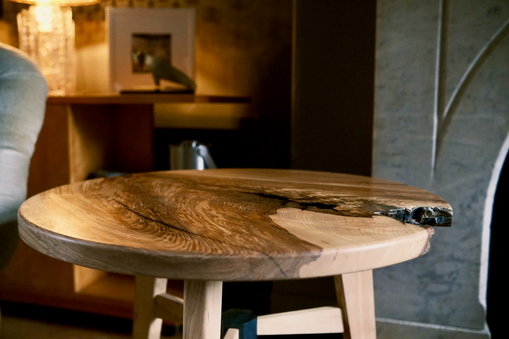 A rustic live-edge ash wooden table with a round top and light-colored legs, shown in a warm, dimly lit interior setting.