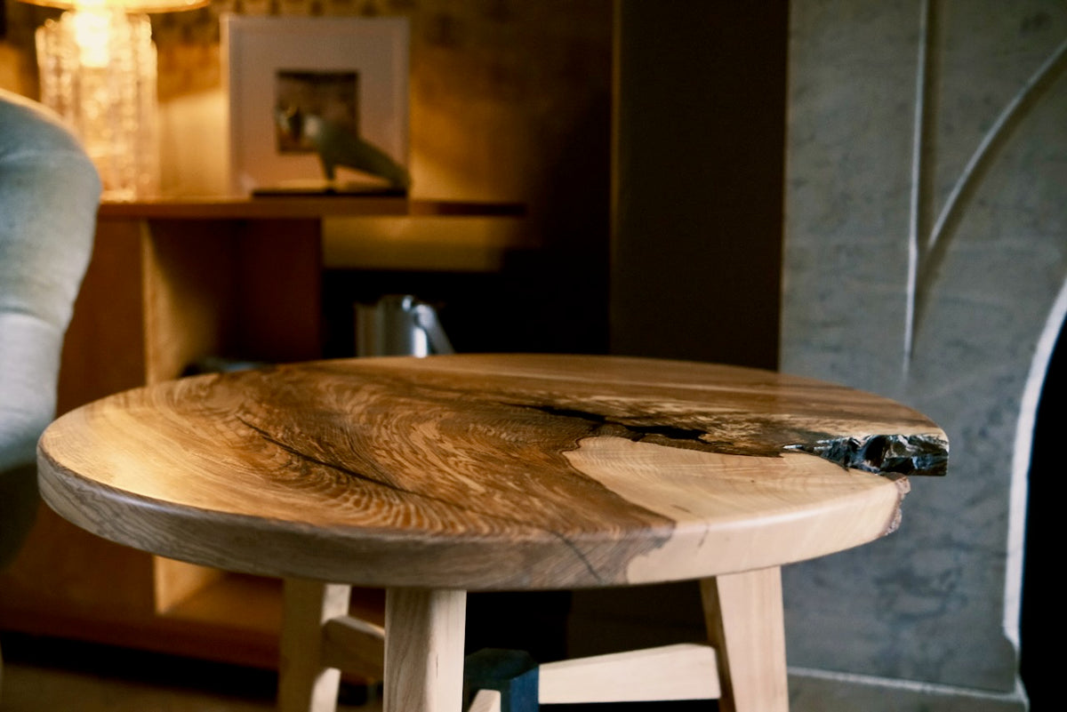 A rustic live-edge ash wooden table with a round top and light-colored legs, shown in a warm, dimly lit interior setting.